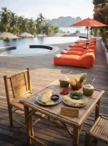 Table en bambou avec petit-déjeuner thaïlandais et noix de coco face à une piscine à débordement et des falaises tropicales, illustrant la liberté financière et le bien-être en Thaïlande.